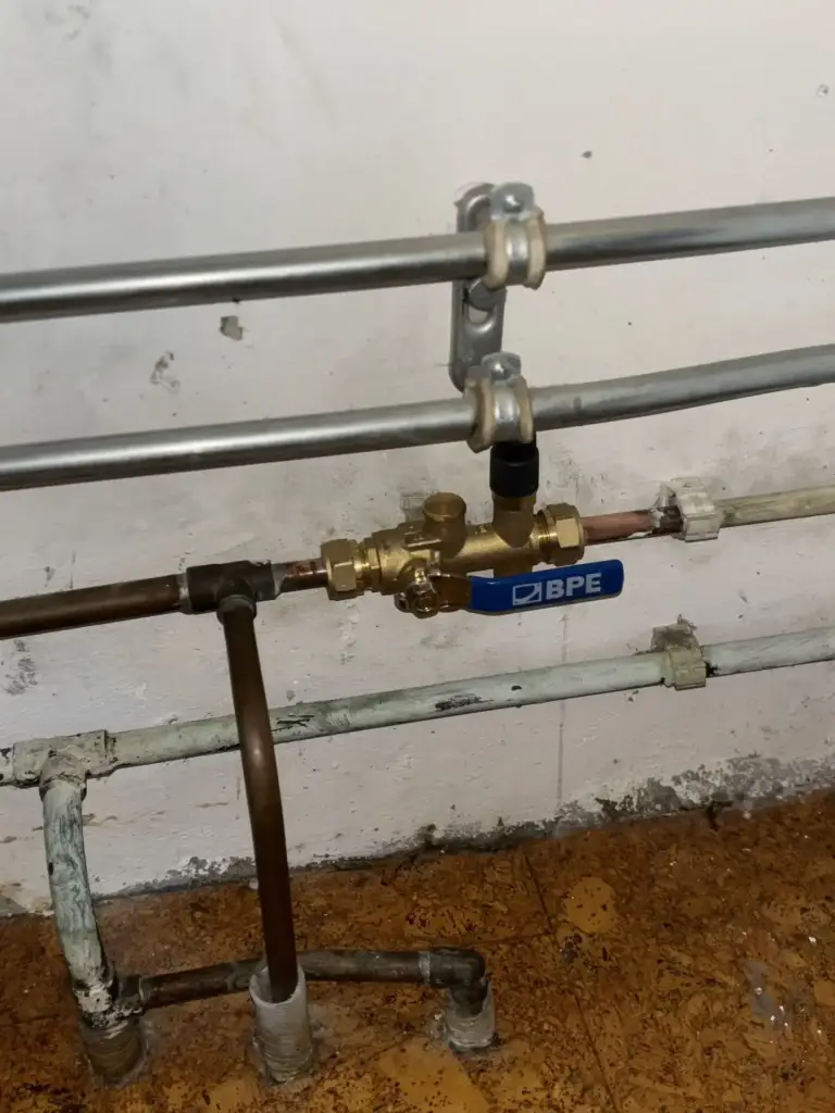 Koperen waterleiding met messing kogelkraan tegen een witte muur.