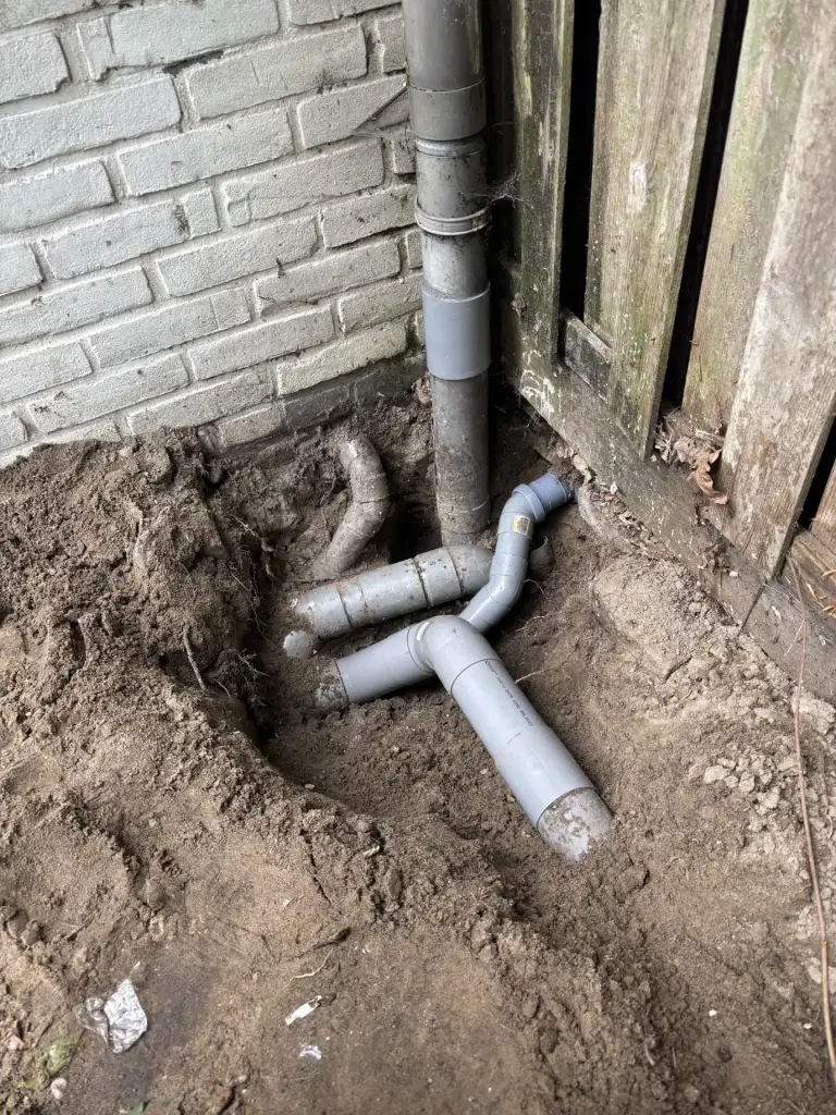 Vrijgegraven grijze PVC-afvoerleidingen naast een regenpijp tegen de gevel.