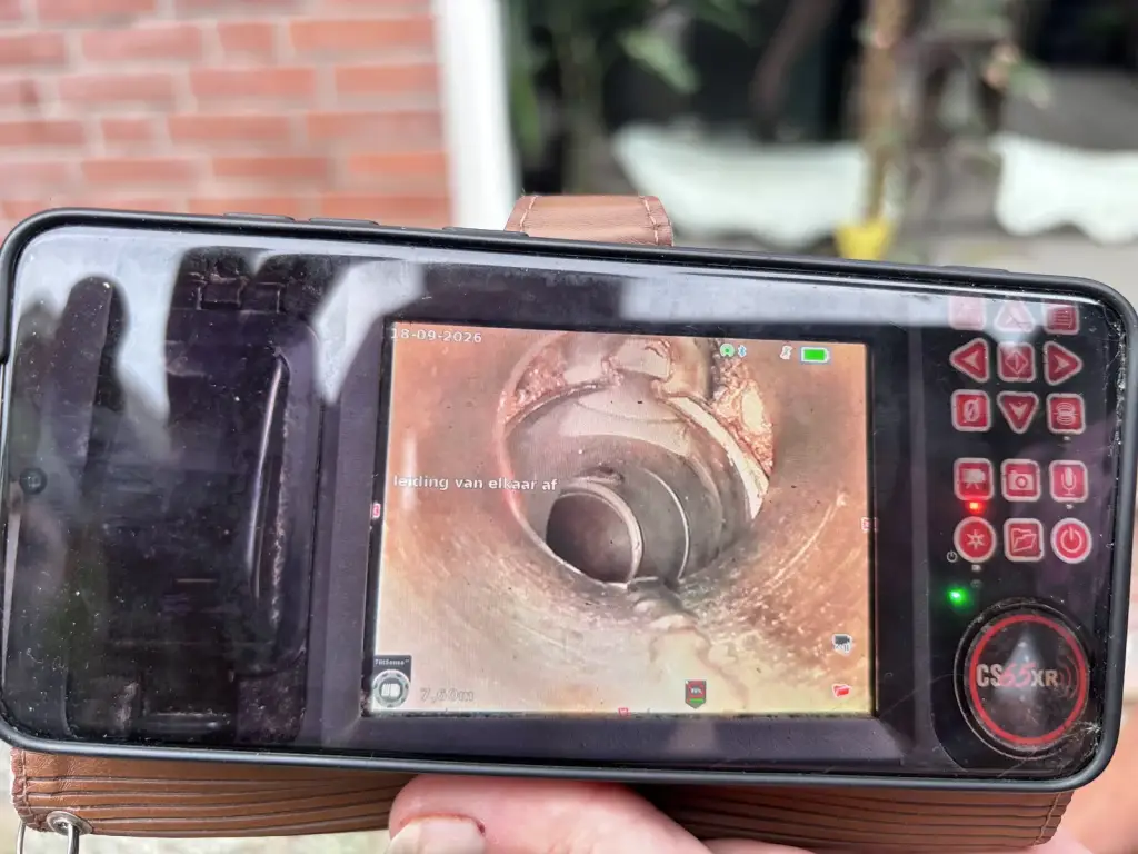Monteur houdt het scherm van een rioolcamera vast; op het scherm is de binnenkant van een afvoerbuis te zien.